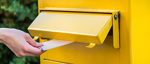 Symbolbild: Ein gelber Briefkasten, in den gerade von einer Hand in Großaufnahme ein Briefumschlag eingeworfen wird