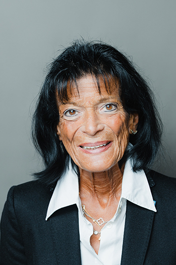 Bildbeschreibung: Porträtfoto von Marianne Frickel, auf dem sie lächelnd in die Kamera blickt. Sie ist 71 Jahre alt, trägt halblange schwarze Haare, eine weiße Bluse und einen schwarzen Blazer.