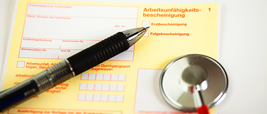 Das Bild zeigt eine Arbeitsunfähigkeitsbescheinigung. Darauf liegt ein Kugelschreiber und ein Stethoskop.