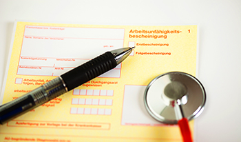 Das Bild zeigt eine Arbeitsunfähigkeitsbescheinigung. Darauf liegt ein Kugelschreiber und ein Stethoskop.
