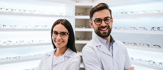 Auf dem Foto sind eine Optikerin und ein Optiker in ihrem Geschäft mit zahlreichen Brillen im Hintergrund zu sehen.